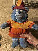 Front view of blue plush bear wearing orange tunic and red hat