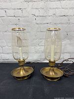 Front view of both brass and glass hurricane table lamps showing bases, glass chimneys, cords, and overall wear