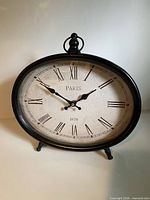 Front view of black metal oval clock with Roman numerals and Paris 1878 text