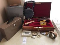 Photo of entire lot showing wood inlay gun case opened with red velvet interior, two metal lock boxes, leather holster, belt with buckles, circular metal canteen with fabric cover and carrying strap, and small boxed accessories on a surface near window.