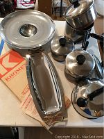 Photo of vintage Revere Ware tea kettle, three pans with lids, extra lid, plus Kromex unique serving tray with wood raised base and original box on table.