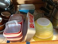 Shelf with four red Pyrex rectangular storage dishes, two have lids, turquoise loaf and divided dish with lids, and stacked yellow mixing bowls.