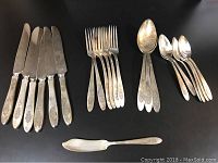 Full set of 6 knives, 6 forks, 3 large serving spoons, 6 small spoons and 1 butter knife arranged on table, showing engraved Bird of Paradise pattern and tarnished condition.