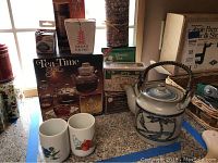 Full view of vintage stoneware teapot, two ceramic tea cups, boxed glass teapot, unopened tea balls, and tea canisters on table.
