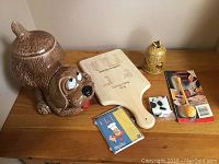 Photo showing McCoy dog cookie jar, beehive honey jar, wooden recipe board, chef magnets, and cow tape measure on wooden table