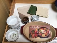 Top view of four white ramekins, two with green leaf design, two small brown and blue ceramic bowls, two square decorative trivets (green and cream), one triangular trivet, and Pampered Chef Crockery Pot recipe book on oval casserole dish.