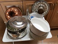Overview of cookware and bakeware including copper pudding mold, CorningWare dishes, stainless steel pieces all stacked on tray