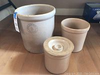 Three beige stoneware crocks arranged on a wooden floor, one with marked brand, one medium sized, and one small crock with lid