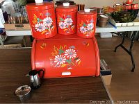 Photo showing the red metal bread box with three matching canisters sitting on top, all decorated with white daisy flowers and colorful leaves.