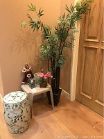 Porcelain painted stool, imitation bamboo plant, handmade ethnic doll, small decor items against wall near wood door.
