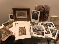 Wide view of a table with a framed religious artwork and multiple loose vintage religious prints spread around it, including black and white and color prints.