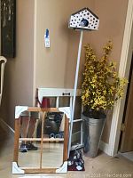 Full view of the birdhouse on a white pole with red and white pattern, tall artificial yellow flower arrangement in a metal bucket, the 19x23 inch wood framed mirror leaning against the wall with corner protectors and small red metal watering can on top of it, next to a white chair.