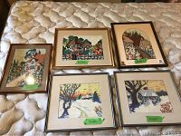 Five framed stitchery pictures laid out on a mattress, showing detailed embroidered rural and home scenes with vibrant colors.