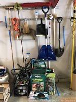 Photo showing wall-mounted garden tools including brooms, shovels, rake, dustpan, and pile of hose connectors and hose sprayer systems in boxes.