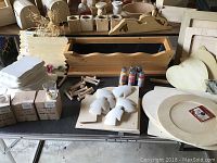 Collection of unfinished wood craft items including planters with scalloped edges, plaques, wooden spindles, and boxed paint bottles arranged on a table.