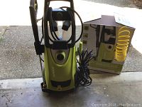 Full view of Sunjoe electric pressure washer with power cord, attached hose, spray gun, and original box in background on concrete floor.