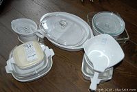 White ceramic Corning Ware serving dishes with glass lids and Gravy Mate gravy boat, various sizes and shapes, arranged on brown wooden surface