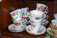 Twelve assorted bone china tea cups stacked on and around matching saucers with floral patterns and gold edging inside a wooden cabinet.