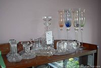 Photo showing crystal decanter, creamer, sugar bowl, four colored stemmed champagne glasses, and assorted small dishes and candle holders on a shelf.