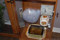 Glass cabinet interior showing various kitchenware items: brie cheese pot with wooden base and lid, ceramic cookie jar with fruit motif, stacked plastic salad bowls, large glass and ceramic serving trays.
