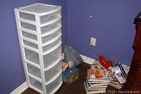 Full view of the Sterilite plastic drawer unit next to a pile of assorted books and items on floor against purple wall.