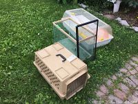 Photo showing glass aquarium, beige pet carrier kennel, and plastic pet cage on grass outdoors
