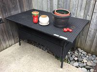 Black vinyl rattan look patio table shown outdoors against a wooden fence, with several small pots and items on top and a rocky garden bed beneath.