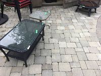 Vinyl rattan look outdoor coffee table with glass top alongside metal framed side table and metal fire pit in the background, showing overall condition and damage points.