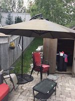 Image showing the full freestanding patio umbrella set up on a patio with a wide fabric canopy that is taupe-colored and supported by black metal frame and base.