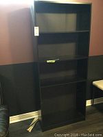 Full front view of the tall black laminate bookcase against the wall, showing four open shelves.