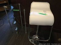 Photo showing the white faux leather cushioned seat of the bar stool and the glass 3-tier corner shelf next to it on the floor.
