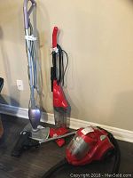 Three vacuum cleaners including one red Dirt Devil canister vacuum, one red Dirt Devil stick vacuum, and one silver/purple Shark wet broom vacuum