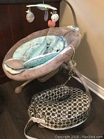 Motorized baby rocking seat with patterned cushion on the floor next to it, showing full seat with padded fabric and attached mobile arm with toys.