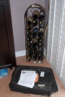 Metal wine rack standing upright against wall, showing multiple wine bottles installed, with Baker Stone BBQ pizza oven on floor in front