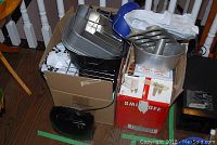 Photo showing assorted metal bakeware like cake pans and cooling racks stacked in cardboard boxes along with some kitchen appliance boxes.