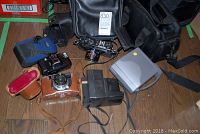 Assorted vintage cameras and cases including Yashica, Polaroid, and a portable DVD speaker case on wooden floor.