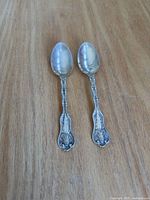 Front view of two ornate sterling teaspoons on wood surface