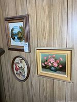 Three framed artworks on wall: two rectangular oil paintings and one oval needlepoint