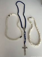 Overall view of three necklaces laid out: blue glass rosary with crucifix, white shell heishi necklace, white shell chip necklace