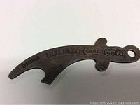 Photo of antique Coca-Cola metal bottle opener showing embossed 'DRINK BOTTLED Coca-Cola' text and overall curved shape with hanging loop.