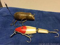 Two vintage fishing lures on blue cloth, one brown animal-shaped with feather tail and three treble hooks, one segmented red and white lure with diving lip and treble hooks.