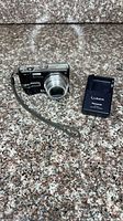 Camera front view with lens extended, wrist strap attached, battery charger alongside