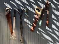 Tools hanging on wall: two hand saws, small leaf rake, forged tongs, garden weeder, pipe wrench