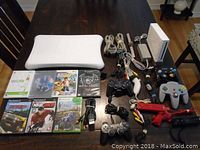 Photo showing Nintendo Wii console, Wii Balance Board, various controllers including Nintendo GameCube, Nintendo 64, Sony PS2/PS3, Wii remotes, red gun attachments, and multiple video game discs arranged on a table.