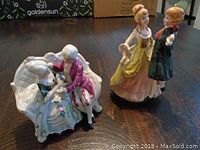 Two porcelain figurines of couples, one seated on a white sofa with the male playing music; the other standing couple interacting.