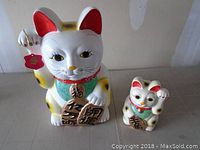 Photo showing both Japanese Maneki Neko lucky cat banks side by side on a surface, illustrating size difference and design details.