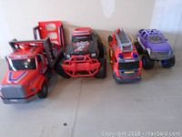 Four toy trucks lined up side by side against wall on a beige floor, including a red car carrier, off-road truck, red fire truck, and purple truck.