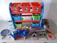 Frontal view of blue wooden storage organizer with multiple plastic bins holding wooden Thomas the Train track pieces and accessories, arranged on and around the organizer including bridges and stations.