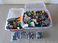 Wide view showing two large plastic containers filled with assorted LEGO blocks and four sealed bags of LEGO pieces in front.