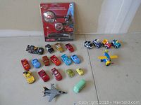 Wide shot showing assortment of small toy cars, trucks, a helicopter, a fighter jet, and Tonka items on a flat surface.